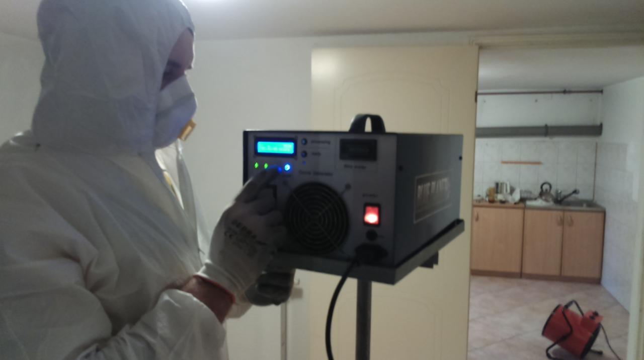 Pracownik w kombinezonie obsługuje ozonator w pomieszczeniu z widoczną kuchnią. Urządzenie na statywie, w tle wentylator. Dezynfekcja po zalaniu.