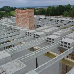 Widok z góry na strop Teriva w trakcie montażu, z częściowo wymurowanym kominem z cegły klinkierowej. Szare bloczki stropowe z otworami, drewniane stemple podtrzymujące.