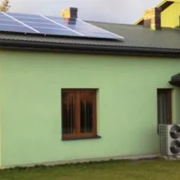 Dom jednorodzinny z panelami fotowoltaicznymi na dachu i pompą ciepła na zewnątrz, otoczony zielonym trawnikiem. Widok z boku, słoneczny dzień.