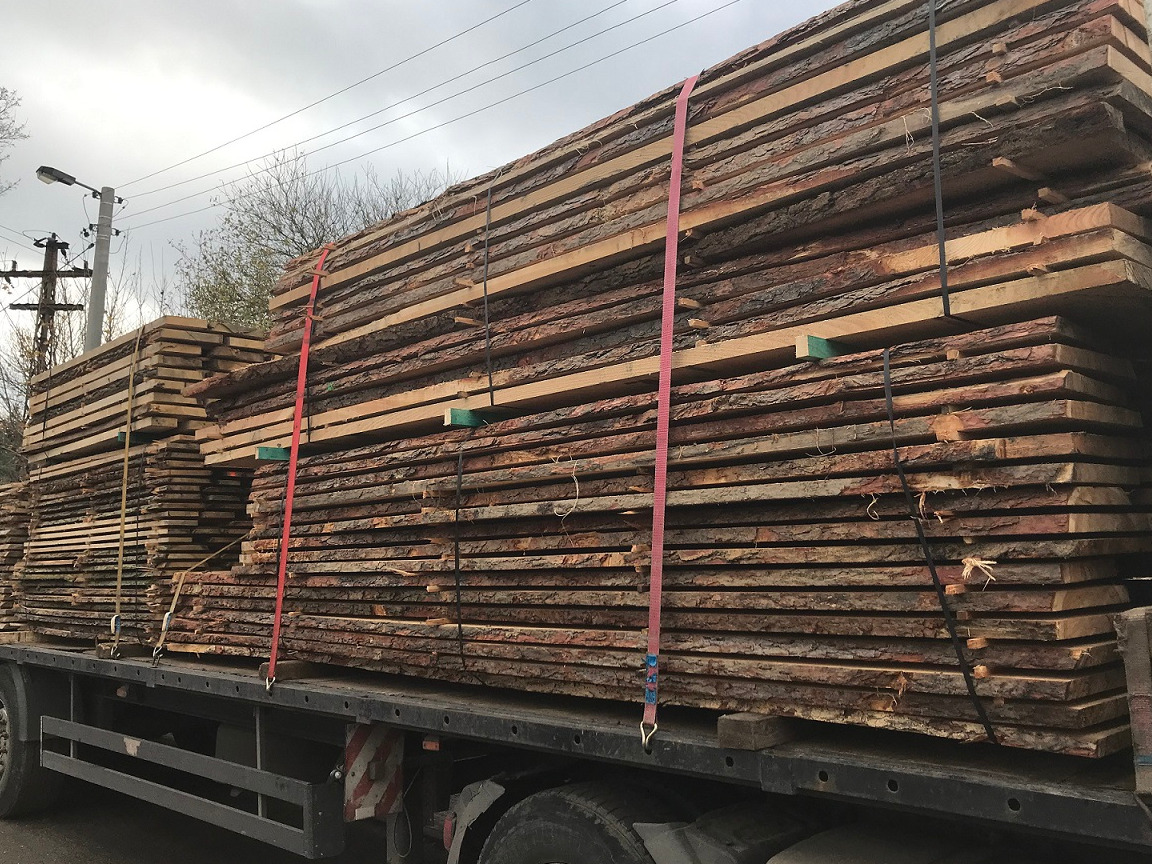 Ciężarówka załadowana stosem świeżo przetartego drewna z korą, zabezpieczonego czerwonymi pasami transportowymi.