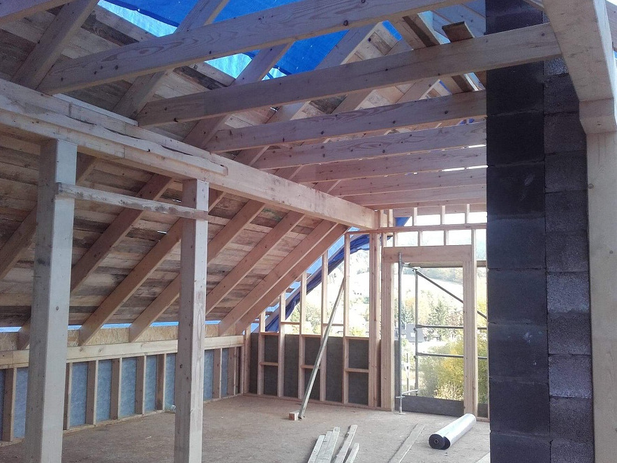 Wnętrze budynku w trakcie budowy, widoczna drewniana konstrukcja dachu i ścian, częściowo ocieplone ściany, niebieska plandeka zabezpieczająca dach, bloczki betonowe przy otworze drzwiowym.