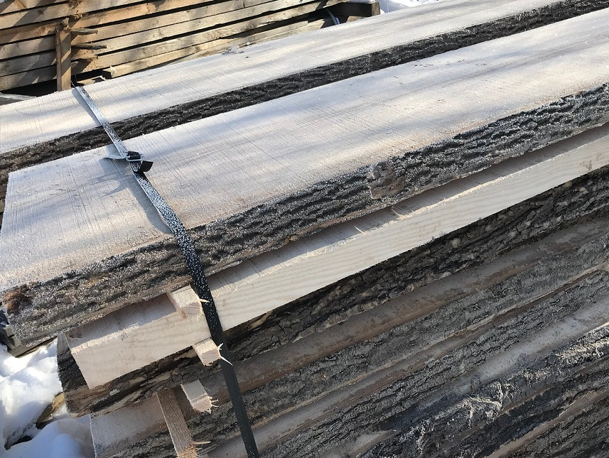 Stos desek z korą, spięty czarną taśmą, widoczny lekki szron na powierzchni, w tle inne deski i śnieg.