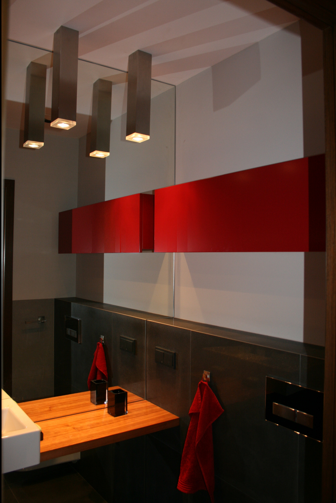 Nowoczesna łazienka z drewnianym blatem, czerwonymi akcentami i podłużnymi lampami sufitowymi odbijającymi się w lustrze.