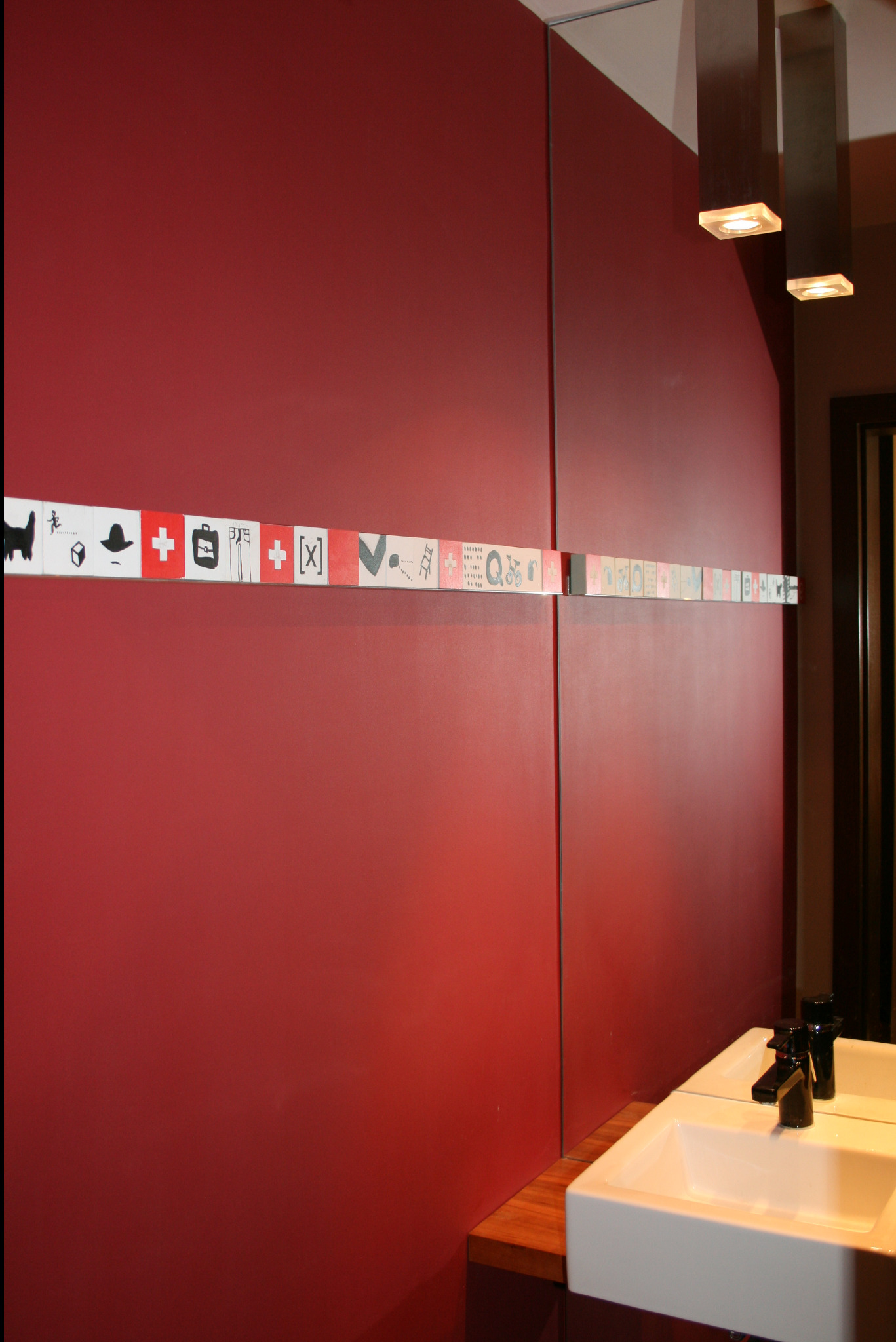 Czerwona ściana w łazience z dekoracyjnym paskiem z ikonami, biała umywalka z czarną baterią, nowoczesne lampy wiszące.