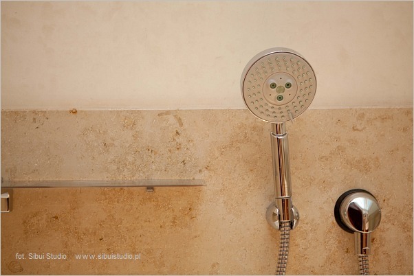 Fragment łazienki z beżową ścianą z kamienia, chromowaną słuchawką prysznicową z zielonymi akcentami i minimalistyczną szklaną półką.