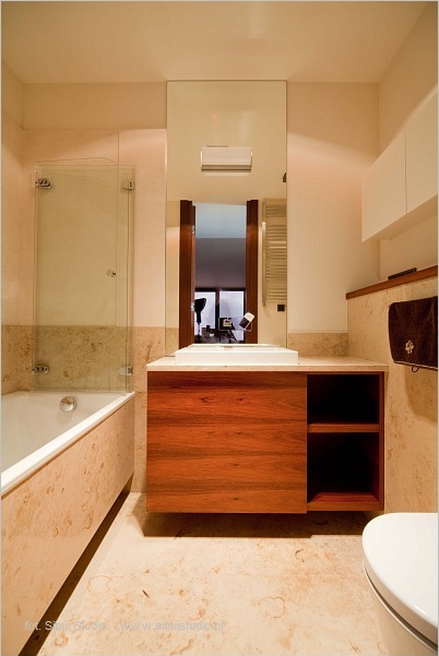 Nowoczesna łazienka z beżowymi płytkami imitującymi marmur, drewnianą szafką pod umywalką z otwartymi półkami i dużym lustrem.