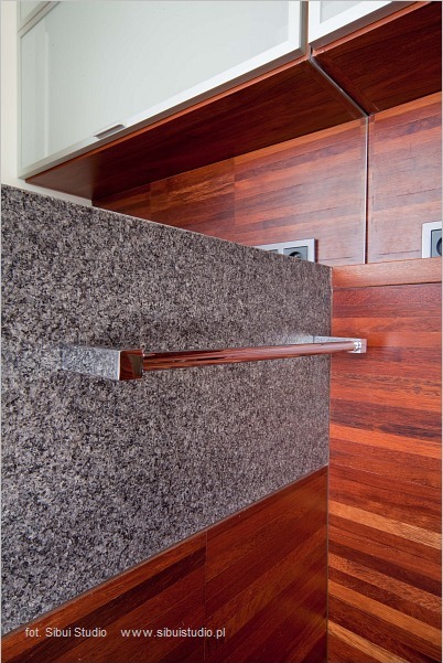 Fragment łazienki z kamienną ścianą, drewnianą podłogą ułożoną w jodełkę i chromowanym wieszakiem na ręczniki. Widoczne gniazdka elektryczne w drewnianej zabudowie.