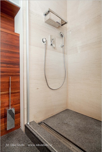 Nowoczesna kabina prysznicowa z deszczownicą, wykończona jasnym trawertynem i ciemnym, grafitowym kamieniem na podłodze z odpływem liniowym. Drewniane elementy wykończeniowe widoczne obok kabiny.