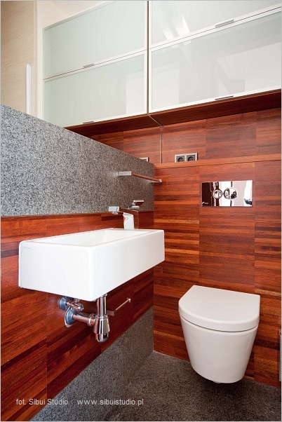 Nowoczesna łazienka z drewnianą okładziną ścienną, prostokątną umywalką nablatową, granitowym blatem i podwieszaną toaletą, widoczne gniazdka elektryczne i chromowana armatura.