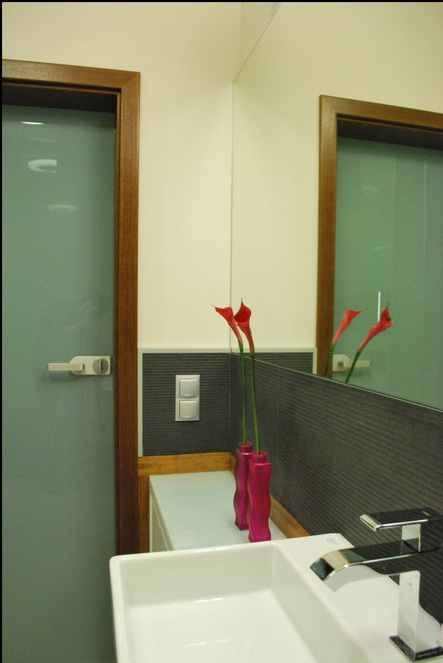 Umywalka ceramiczna z chromowaną baterią, dwie różowe wazy z kaliami na tle szarej ściany z teksturą i białym włącznikiem światła w nowoczesnej łazience z lustrem i drzwiami z drewnianą ramą.