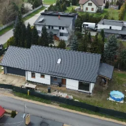 Widok z góry na dom jednorodzinny z ciemnoszarą dachówką, z oknem dachowym, ogrodzony zieloną siatką, z widocznymi elementami placu budowy i otoczeniem zieleni, w tle inne budynki.