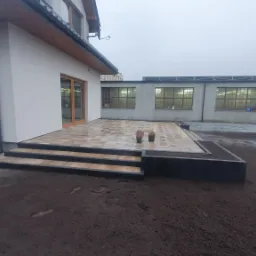 Nowoczesny taras z płytek na trzech poziomach, z ciemnymi obrzeżami i donicami, przylegający do białego budynku z drewnianą stolarką okienną, w tle budynek przemysłowy.