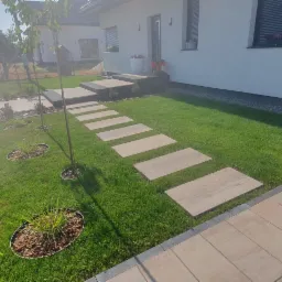 Ogród przydomowy z trawnikiem, ścieżką z płyt kamiennych i nowoczesnym ogrodzeniem panelowym.