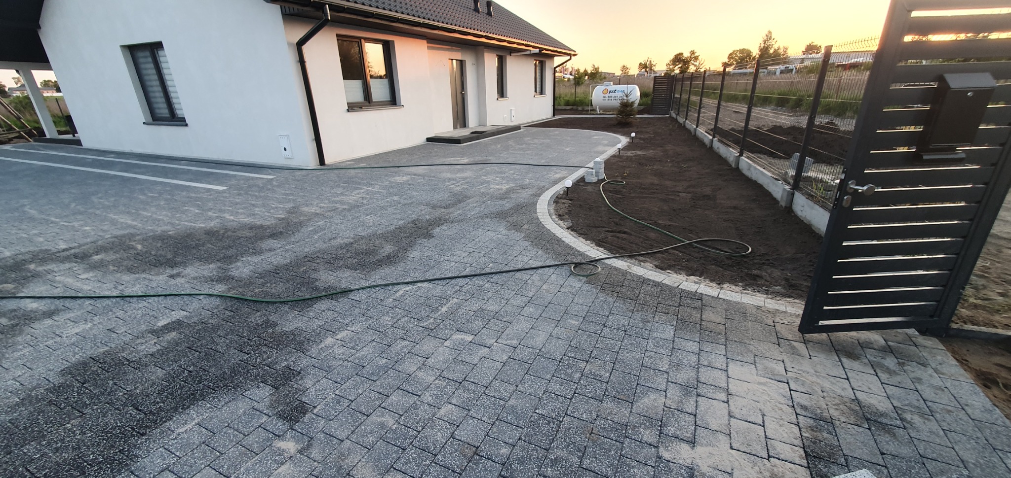 Nowoczesny podjazd z szarej kostki brukowej prowadzący do domu jednorodzinnego z białymi ścianami, obrzeża z jasnej kostki, świeżo przygotowany teren pod trawnik, widoczna brama wjazdowa i zbiornik...