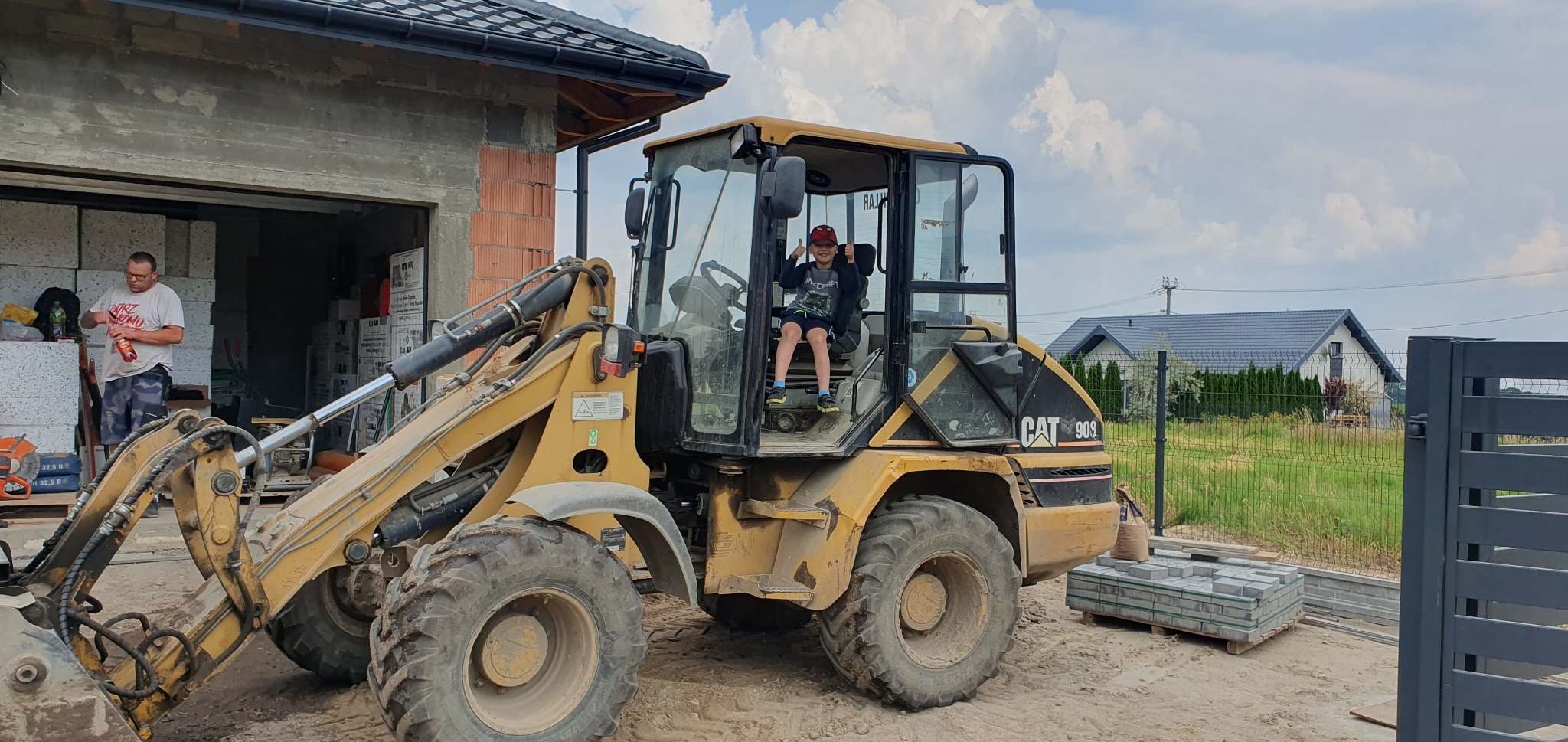 Chłopiec siedzi w kabinie żółtej ładowarki CAT 908 na placu budowy, w tle niedokończony budynek i ułożona kostka brukowa, obok stoi mężczyzna.