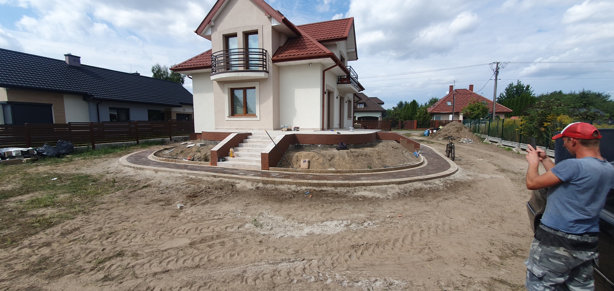 Układanie kostki brukowej wokół nowego domu z balkonem, widoczny etap prac ziemnych i częściowo ułożona ścieżka z kostki. Mężczyzna w czerwonej czapce robi zdjęcie telefonem.
