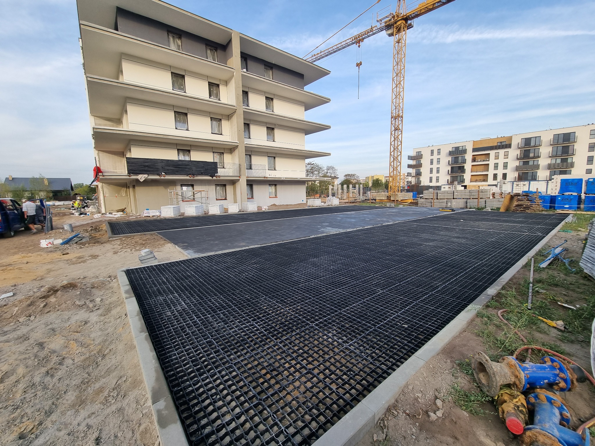 Przygotowanie podłoża pod parking z wykorzystaniem plastikowej kratki, widoczny budynek wielorodzinny w trakcie budowy oraz żuraw wieżowy.