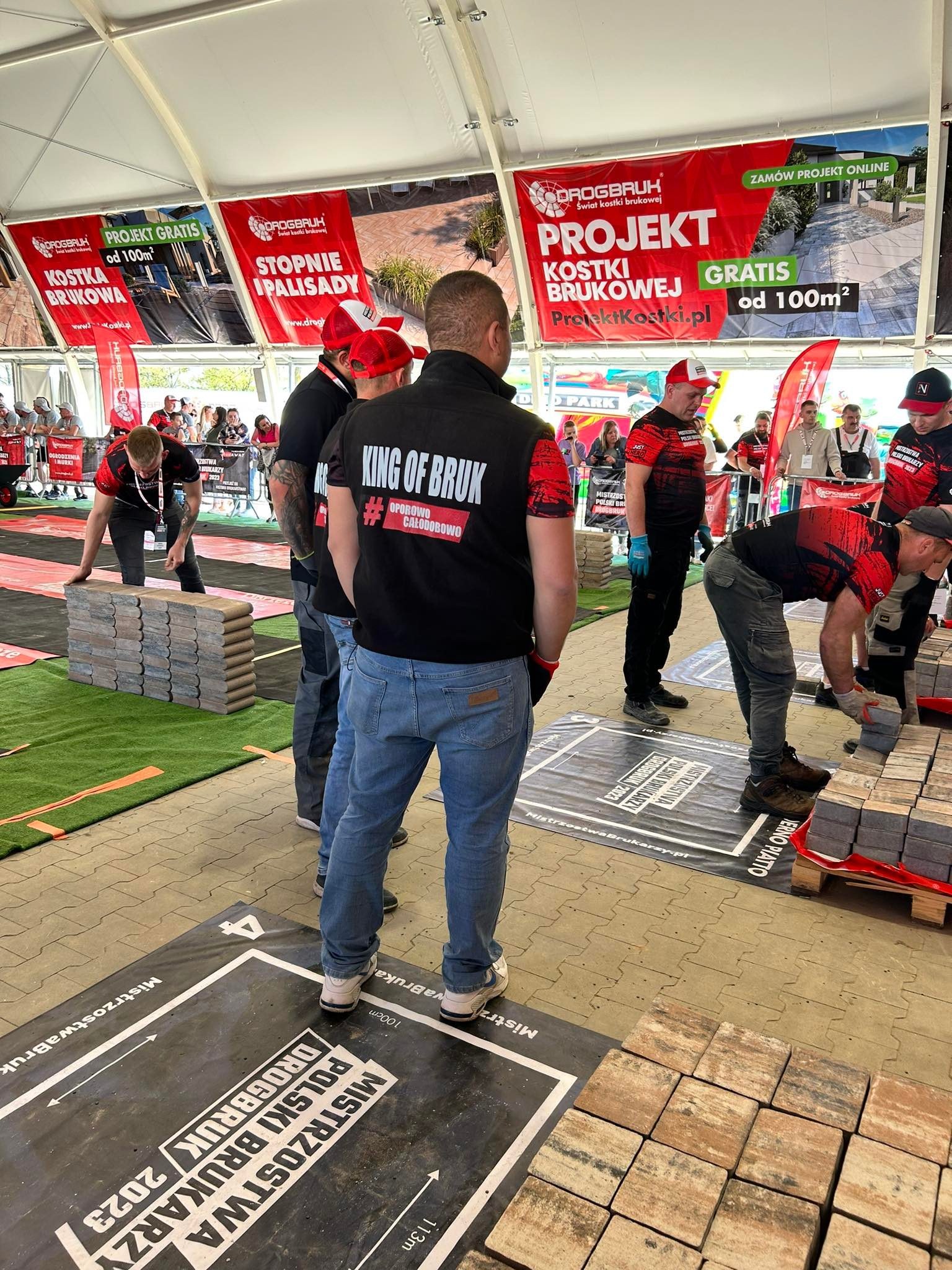 Zawody brukarzy: uczestnicy układają kostkę brukową na wyznaczonych stanowiskach, widoczne bannery reklamowe z ofertą projektów kostki brukowej gratis powyżej 100m2, hasła 'King of bruk'...
