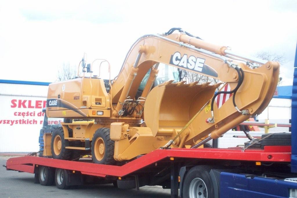 Żółta koparka CASE na czerwonej platformie lawety, widok z boku, w tle budynek z napisem 'SKLEP'.