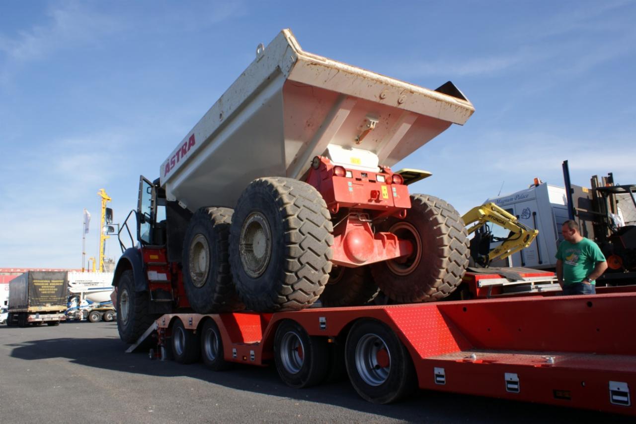 Ciężki wywrotka Astra z ogromnymi oponami załadowana na czerwoną niskopodwoziową lawetę. Widoczny fragment kabiny ciągnika oraz pracownik w tle.