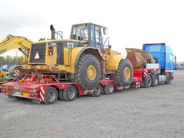 Transport niskopodwoziowy: żółta ładowarka CAT 980G na czerwonej platformie, ciągnięta przez niebieską ciężarówkę. Widoczne łańcuchy zabezpieczające ładunek.