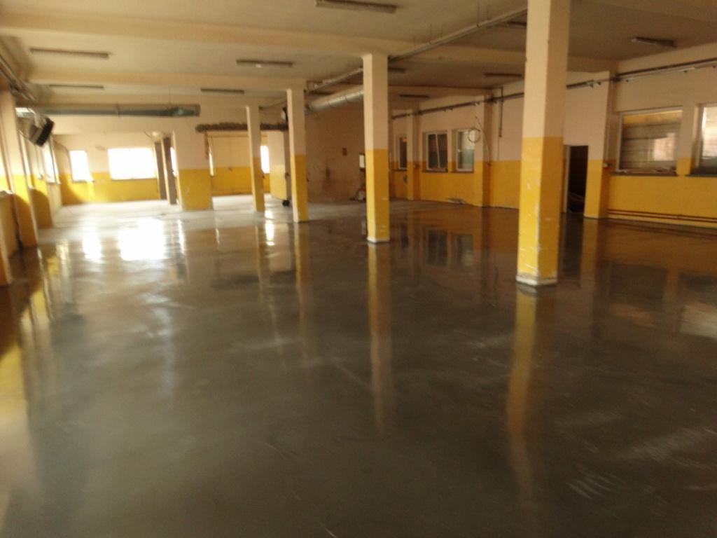 Błyszcząca, szara posadzka w przestronnym, industrialnym wnętrzu z żółto-białymi kolumnami i oknami. Widoczne refleksy światła na gładkiej powierzchni.