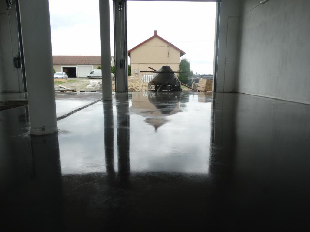 Błyszcząca, ciemna posadzka żywiczna w hali, odbicie budynku w połysku, widoczne słupy konstrukcyjne. Gładka powierzchnia, surowy, industrialny charakter.