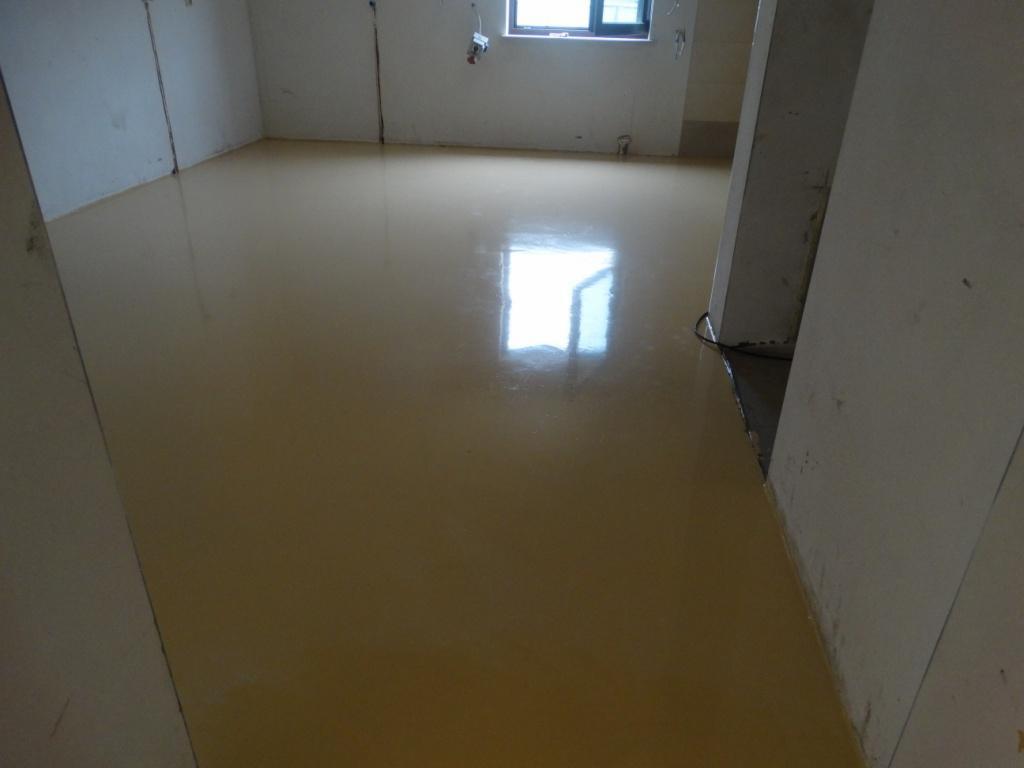 Jasna, świeżo wylana posadzka w surowym wnętrzu. Gładka powierzchnia odbija światło z okna. Widoczne ślady po pracach wykończeniowych na ścianach.