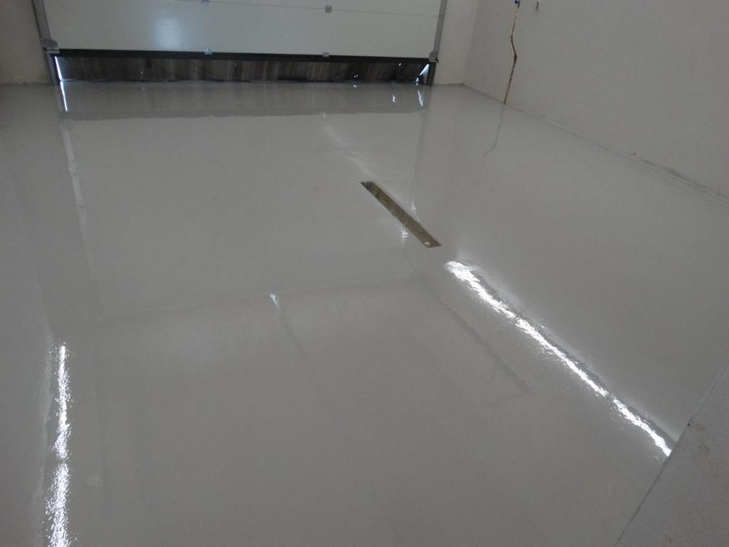 Gładka, szara posadzka z odpływem liniowym w garażu. Widoczna brama garażowa i pęknięcie na ścianie. Nowoczesne wykończenie podłogi.