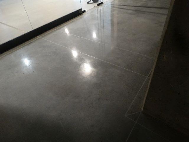 Szara, błyszcząca posadzka z mikrocementu z widocznymi liniami podziału, odbijająca światło. Fragment ściany w tle.