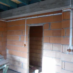 Wewnętrzna instalacja elektryczna w surowym budynku z cegły: białe kable prowadzone po ścianach i pod sufitem, widoczne puszki instalacyjne i fragment rusztowania.