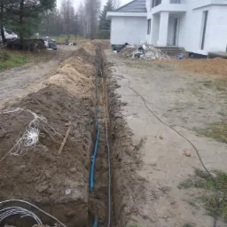 Wykop z ułożonymi przewodami (niebieski i szary) oraz kablem w ziemi, widoczny klucz nastawny, w tle biały dom w trakcie budowy i drzewa.