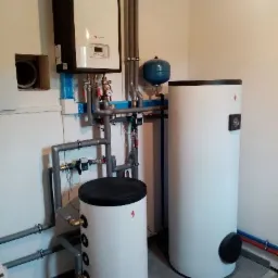 Nowoczesna instalacja grzewcza w kotłowni: biały zbiornik akumulacyjny, wiszący piec z czarną obudową i licznymi rurami z zaworami.