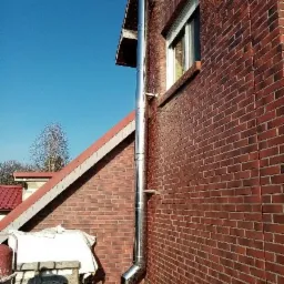 Zewnętrzny, chromowany komin spalinowy przytwierdzony do ceglanej ściany budynku, widoczny stos drewna i fragment dachu pod błękitnym niebem.