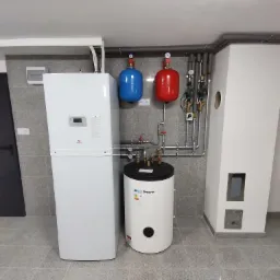Nowoczesna instalacja grzewcza z białą pompą ciepła, cylindrycznym zbiornikiem buforowym Kospel, czerwonym i niebieskim naczyniem przeponowym oraz rurami z zaworami w kotłowni wyłożonej szarymi...