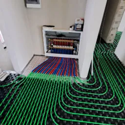 Rozłożone, zielone rury ogrzewania podłogowego na czarnej macie systemowej, z rozdzielaczem z czerwonymi i niebieskimi rurami w tle, widok z góry.