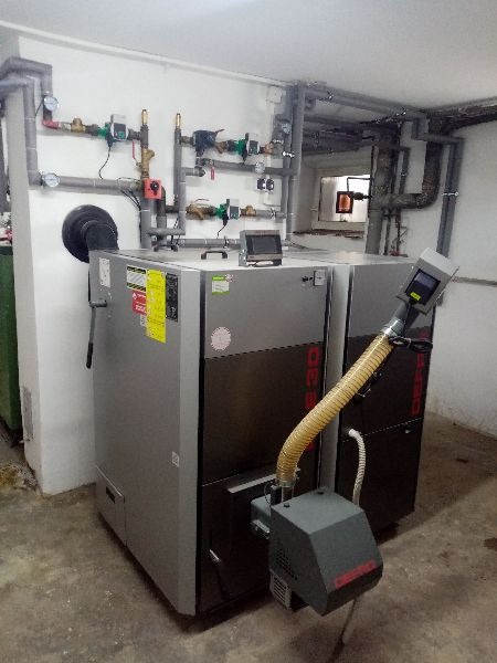 Szary piec na paliwo stałe z podajnikiem i sterownikiem, z widocznymi rurami instalacji CO w tle, w pomieszczeniu kotłowni.