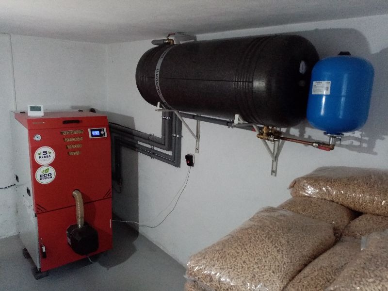 Czerwony piec na pellet z zasobnikiem, zbiornikiem akumulacyjnym i niebieskim naczyniem wzbiorczym w kotłowni, obok worki z pelletem.