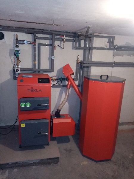 Czerwony piec na paliwo stałe marki Tekla z podajnikiem ślimakowym i zasobnikiem na ekogroszek, zainstalowany w kotłowni z widoczną instalacją grzewczą na ścianie.