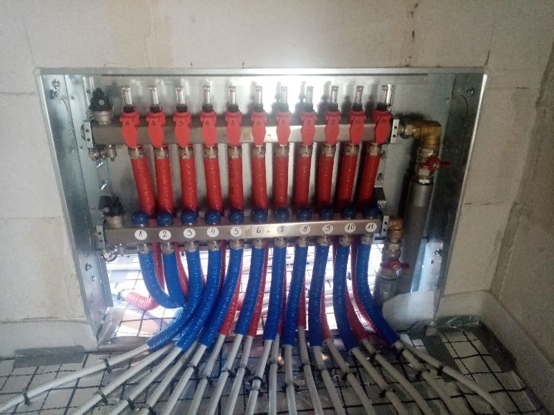 Rozdzielacz ogrzewania podłogowego z czerwono-niebieskimi rurami w izolacji, ułożonymi na siatce montażowej w surowym pomieszczeniu.