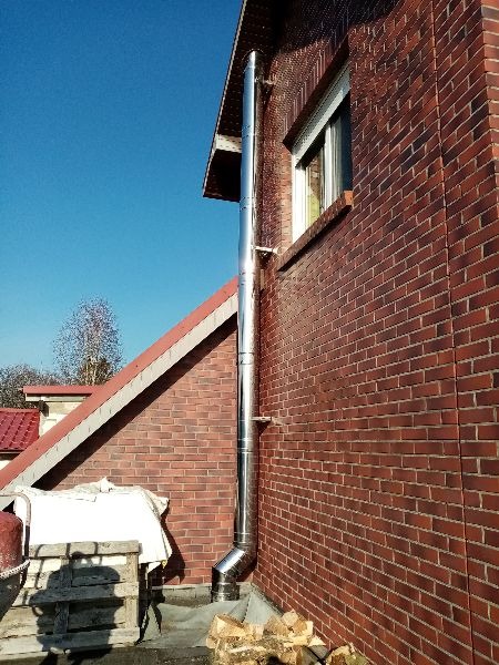 Zewnętrzny, chromowany komin spalinowy przytwierdzony do ceglanej ściany budynku, widoczny stos drewna i fragment dachu pod błękitnym niebem.