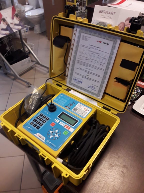 Żółta walizka z miernikiem ZK 100 PRO i certyfikatem kalibracji Labtronik, widoczne kable, tło z armaturą łazienkową i materiałami reklamowymi.