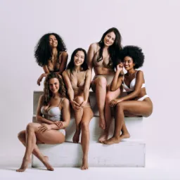 Pięć uśmiechniętych kobiet o różnej karnacji i typach urody prezentuje bieliznę w odcieniach beżu i bieli, siedząc na białych, geometrycznych blokach w jasnym studiu.