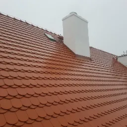 Nowy dach z czerwonej dachówki karpiówki ułożonej łuskowo, z widocznym kominem i oknem dachowym, sfotografowany w pochmurny dzień.