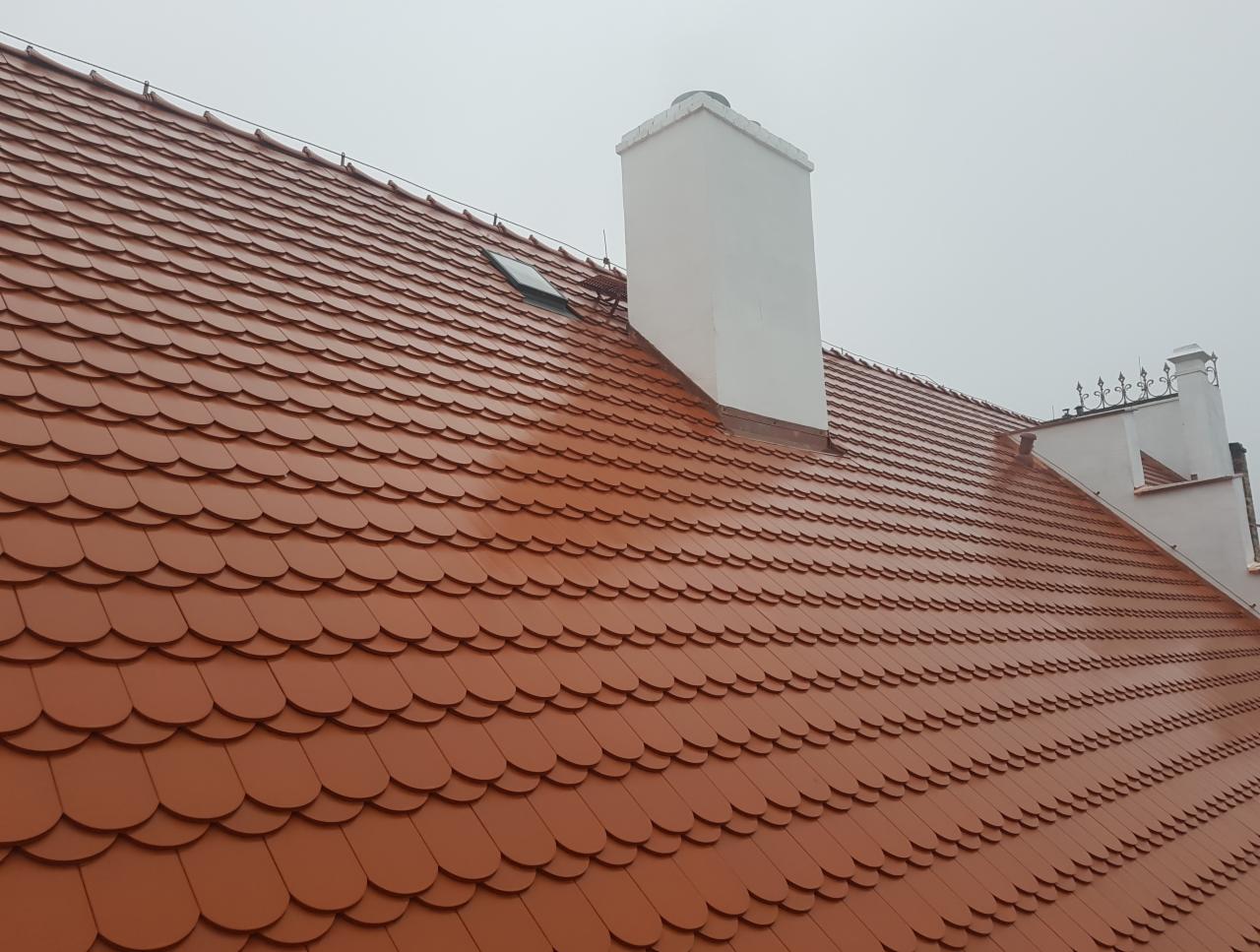Nowy dach z czerwonej dachówki karpiówki ułożonej łuskowo, z widocznym kominem i oknem dachowym, sfotografowany w pochmurny dzień.