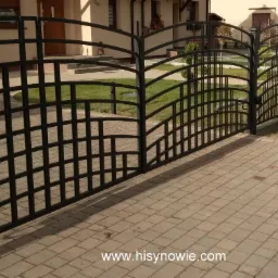 Czarna, metalowa brama wjazdowa i ogrodzenie posesji z ozdobnymi, łukowatymi elementami, widok z zewnątrz na kostkę brukową i budynek mieszkalny w tle.