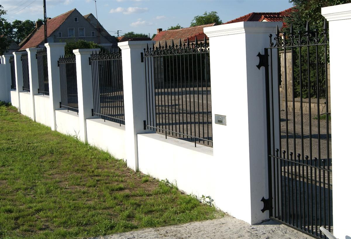Nowoczesne ogrodzenie z białymi słupkami i czarnymi, kutymi przęsłami oraz bramą, widoczne na tle domu z czerwonym dachem i zieleni ogrodu.