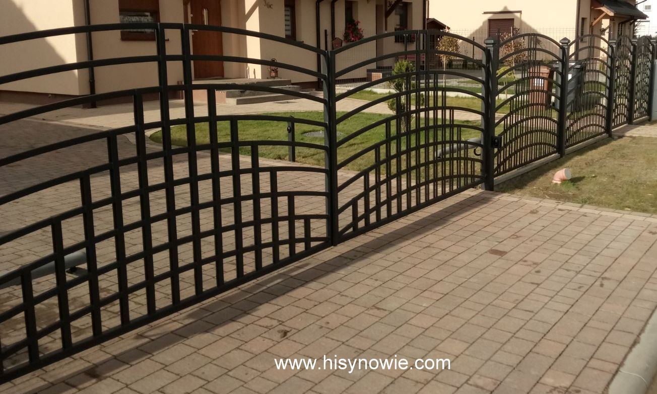 Czarna, metalowa brama wjazdowa i ogrodzenie posesji z ozdobnymi, łukowatymi elementami, widok z zewnątrz na kostkę brukową i budynek mieszkalny w tle.