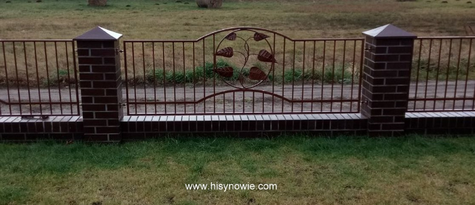 Metalowe ogrodzenie z dekoracją liści, wsparte na niskim murku z cegły, zakończonymi brązowymi daszkami w kształcie piramid, na tle trawnika i polnej drogi.