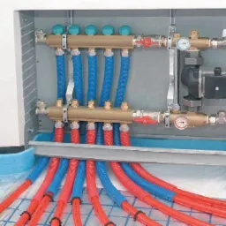 Rozdzielacz ogrzewania podłogowego z czerwonymi i niebieskimi rurami, widoczne manometry i zawory, zamontowany w szafce.